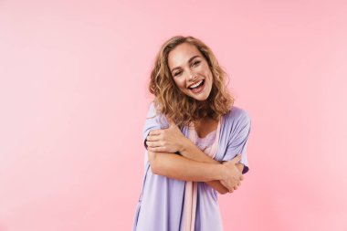 Pembe arka planda kendini izole edip gülümseyen, gülümseyen ve sarılan zarif pijamalı genç bir kadın resmi.