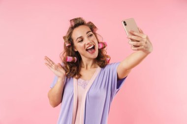 Pembe arka planda izole edilmiş cep telefonundan selfie çekerken bigudili genç bir kadının el sallaması.