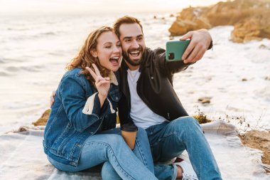 Deniz kenarında yürürken cep telefonuyla selfie çeken güzel beyaz bir çift resmi.