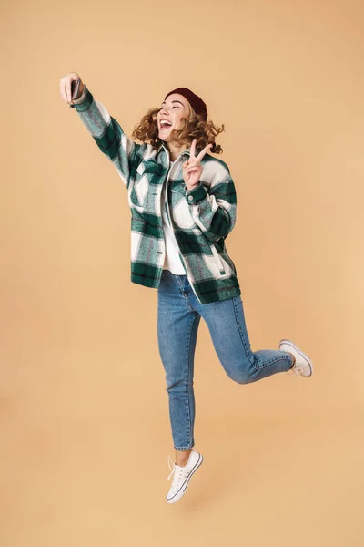 照片中戴着针织帽子的漂亮快乐的女人 一边自拍一边为和平标志 一边跳过米色的背景 — 图库照片
