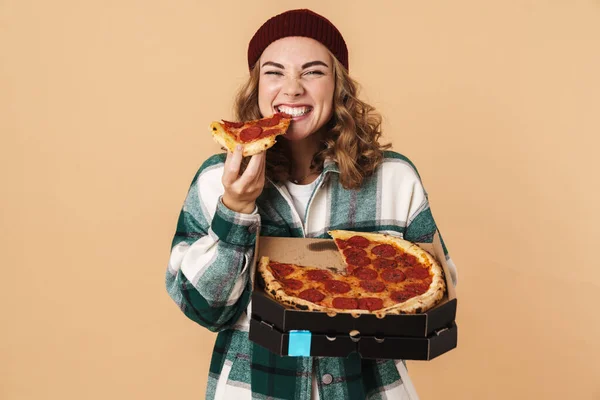 照片上的漂亮的快乐女人戴着针织帽子微笑着 在镜头前吃着比萨饼 背景是米黄色的 — 图库照片