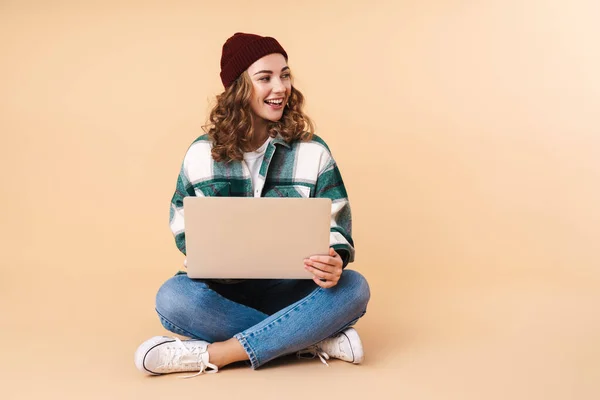 照片中漂亮的快乐女人笑着坐在地板上 用笔记本电脑与米色背景隔离 — 图库照片