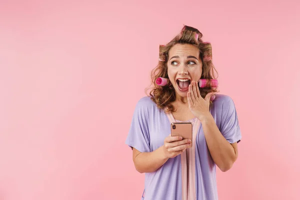 若い素敵な女性の画像でカーラー驚きを表現し ピンクの背景に隔離された携帯電話を使用して — ストック写真