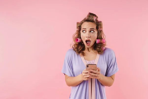 驚きを表現し ピンクの背景に隔離された携帯電話を使用してカーラーで若い怖がっている女性のイメージ — ストック写真