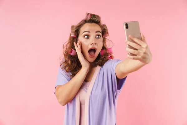 若いです驚きの女性のイメージでカーラー取りますセルフィーオン携帯電話絶縁上のピンクの背景 — ストック写真