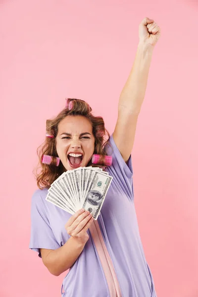在粉红的背景下 一个漂亮而快乐的女人在卷发上做得意洋洋的手势 手里拿着钱 — 图库照片