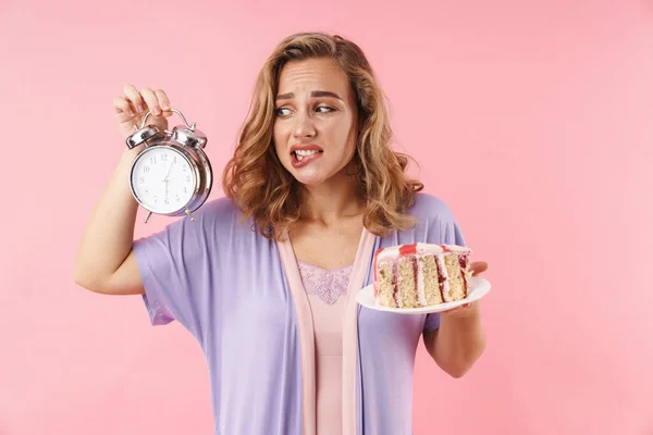 Image of beautiful confused woman in curlers holding alarm and cake isolated over pink background