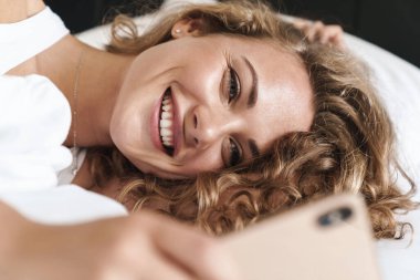 Yatakta yastığa uzanmış, cep telefonu kullanan mutlu, güzel, sarışın bir kadının fotoğrafını çek.