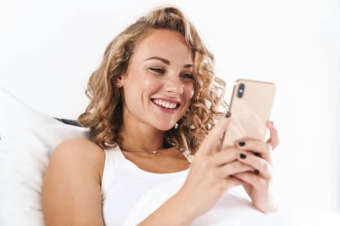 Yatakta yastığa uzanmış, cep telefonu kullanan güzel, genç, sarışın bir kadının fotoğrafını çek.