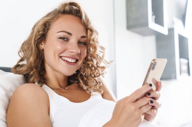 Yatakta yastığa uzanmış, cep telefonu kullanan güzel, genç, sarışın bir kadının fotoğrafını çek.