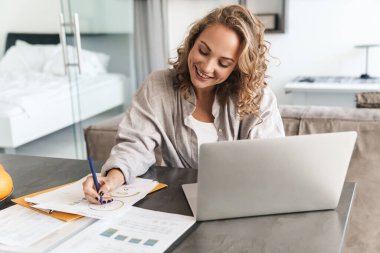 Mutlu, genç, güzel, sarışın bir kadın evde oturmuş dizüstü bilgisayarda çalışıyor, not alıyor.