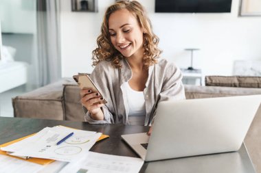 Evdeki masada oturan, dizüstü bilgisayarda çalışan, elinde cep telefonu olan mutlu sarışın kadın.