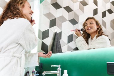 Bornoz giymiş neşeli genç bir kız aynanın karşısında durmuş, makyajını yapıyor.