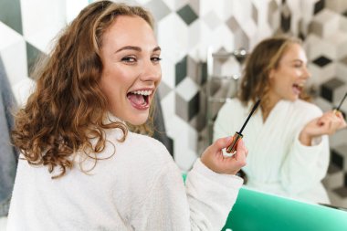 Sabahlık giymiş güzel sarışın bir kadının banyoda duştan sonra aynayla makyaj yaparken görüntüsü.