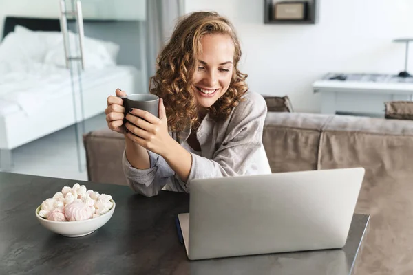 幸せな美しい若いブロンドの髪の女性は 自宅でテーブルに座って ラップトップコンピュータで作業し マグカップを保持 — ストック写真