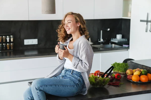 Smiling young woman wearing casual clothes having cup of coffee while sitting on a kitchen table