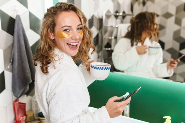 Image of young beautiful blond woman with eye patches holding cellphone and coffee cup in bathroom