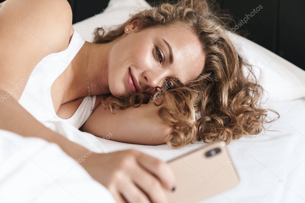 Close up of a smiling beautiful young blonde haired woman laying on a pillow in bed, using mobile phone
