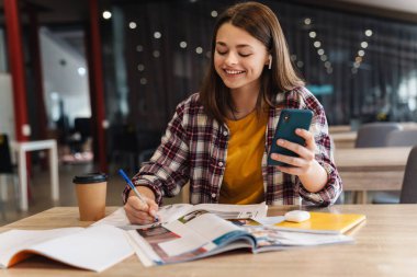 Üniversite kütüphanesinde ödev yaparken kablosuz kulaklık ve cep telefonu kullanan mutlu bir kız resmi