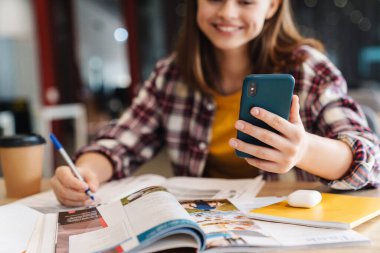 Kolej kütüphanesinde ödevlerini yaparken cep telefonu kullanan neşeli güzel bir kız görüntüsü
