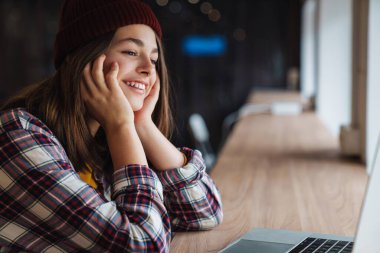 Kolej kütüphanesinde otururken şapkalı, gülümseyen ve dizüstü bilgisayar kullanan neşeli beyaz kız görüntüsü