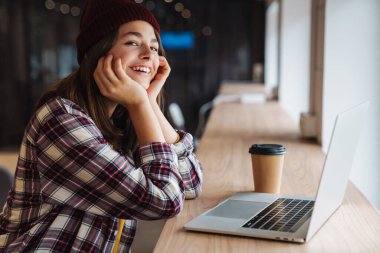 Şapkalı, kahve içen ve kolej kütüphanesinde otururken dizüstü bilgisayar kullanan neşeli beyaz kız görüntüsü.