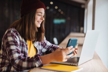 Kolej kütüphanesinde otururken dizüstü bilgisayar ve cep telefonu kullanan şapkalı neşeli beyaz kız görüntüsü