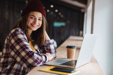 Kolej kütüphanesinde otururken şapkalı, gülümseyen ve dizüstü bilgisayar kullanan neşeli beyaz kız görüntüsü