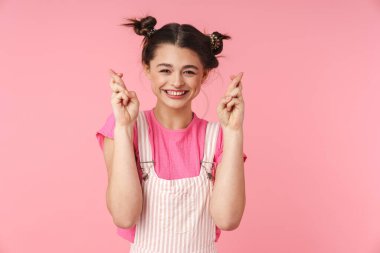 Burun halkalı komik bir kızın fotoğrafı şans dilemek için parmaklarını çapraz tutuyor ve pembe arka planda soyutlanmış bir şekilde gülümsüyor.