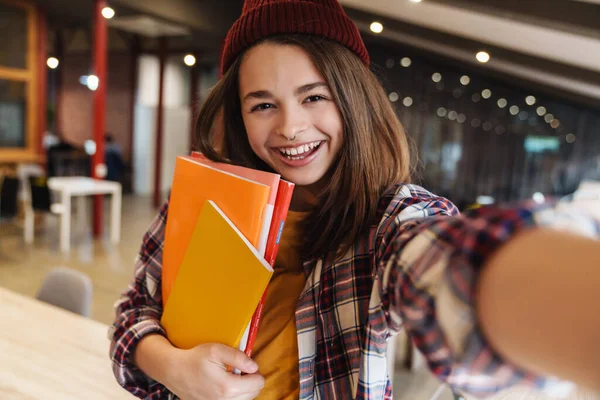 楽しいですCaucasian女の子のイメージで帽子笑顔と取ります自画撮りしながら 大学図書館で運動本を保持 — ストック写真