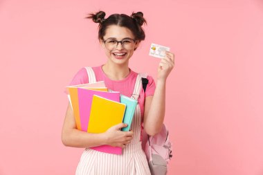 Photo of laughing nice girl in eyeglasses holding exercise books and credit card isolated over pink background clipart
