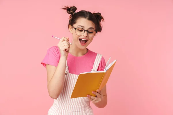 Zdjęcie Podekscytowanej Uroczej Dziewczyny Okularach Piszącej Zeszycie Ćwiczeń Różowym Tle — Zdjęcie stockowe