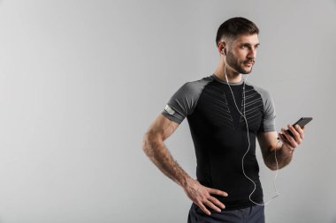 Yakışıklı, güçlü bir sporcunun görüntüsü. Gri duvar arkaplanından izole edilmiş. Cep telefonuyla kulaklıklı müzik dinliyor..