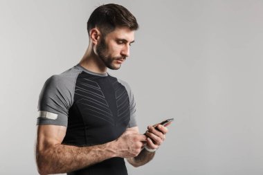 Gri arka planda izole edilmiş, spor kıyafetli, poz veren ve cep telefonu kullanan genç bir adam.