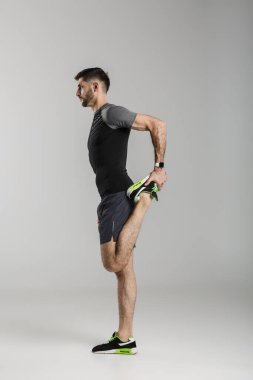 Spor kıyafetli genç bir adamın, gri arka planda tek başına çalışırken bacağını esnetmesi.