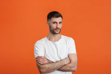 Sade beyaz tişört giyen, elleri turuncu arka planda izole bir şekilde kameraya bakan genç sakallı bir adam.