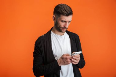 Sade kıyafetler giyen sakallı bir adamın portakal rengi arka planda cep telefonu taşıdığı görüntüsü.