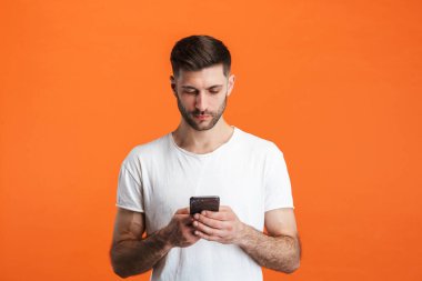 Sade kıyafetler giyen sakallı bir adamın portakal rengi arka planda cep telefonu taşıdığı görüntüsü.