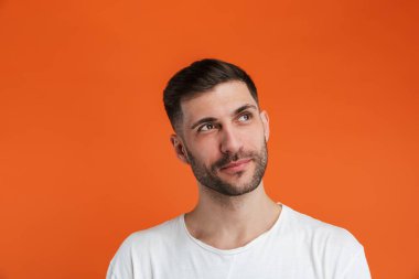 Temel beyaz tişört giyen, gülümseyen ve telif alanına bakan sakallı genç bir adamın portakal rengi arka planda izole edilmiş görüntüsü.