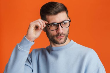 Gözlüklü genç sakallı yakışıklı bir adam portakal rengi arka planda izole edilmiş kameraya poz veriyor.