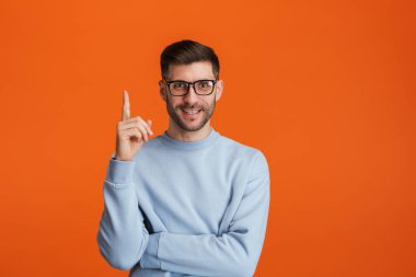 Gözlüklü, sakallı, yakışıklı, gülümseyen ve parmağını turuncu arka planda yukarıya doğru gösteren genç bir adam.
