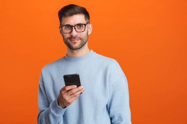 Gözlük takan sakallı yakışıklı bir adamın portakal rengi arka planda izole edilmiş cep telefonu görüntüsü.