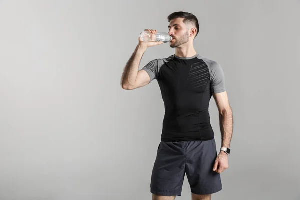 在灰色背景下工作时穿着运动服喝水的年轻强壮男子的形象 — 图库照片