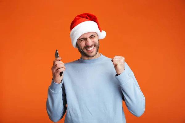 크리스마스 모자를 남자가 핸드폰을 주황색 고립되어 승자의 제스처를 만들고 — 스톡 사진