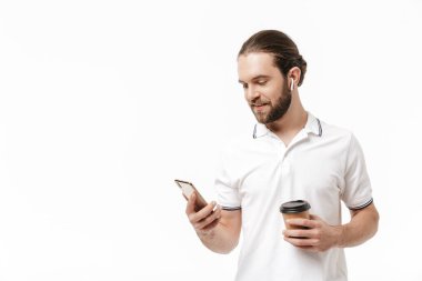 Genç, mutlu ve sakallı bir adamın fotoğrafı beyaz duvar arkasında izole bir şekilde poz veriyor cep telefonu kullanıyor kulaklıklı müzik dinliyor ve kahve içiyor..