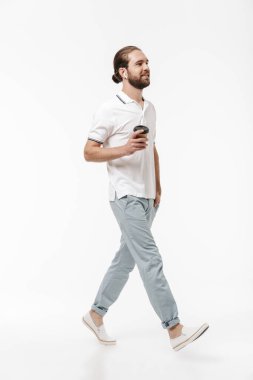 Genç, neşeli, yakışıklı, sakallı bir adamın fotoğrafı. Beyaz duvarın üzerinden izole edilmiş bir şekilde yürüyor. Kulaklıklı, kahve içen müzik dinliyor..