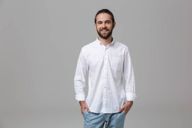 Genç, mutlu, iyimser, yakışıklı, sakallı bir adamın fotoğrafı. Gri duvar arkasında izole bir şekilde poz veriyor..