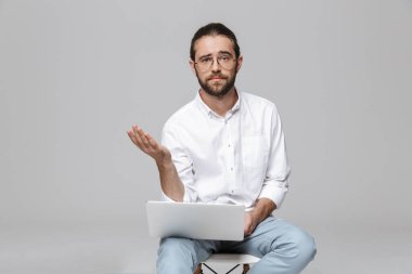 Kafası karışmış, sakallı, gri duvar arkasında dizüstü bilgisayar kullanarak gözlük takan genç bir adamın görüntüsü..