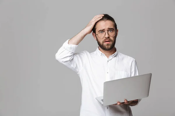 照片中 一个年轻而困惑的英俊大胡子男子 戴着眼镜 用手提电脑在灰色的墙壁上独立地摆姿势 — 图库照片