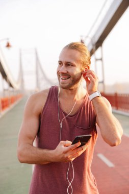 Yakışıklı gülümseyen sporcu, köprüde kablosuz kulaklıkla müzik dinliyor, elinde cep telefonu tutuyor.